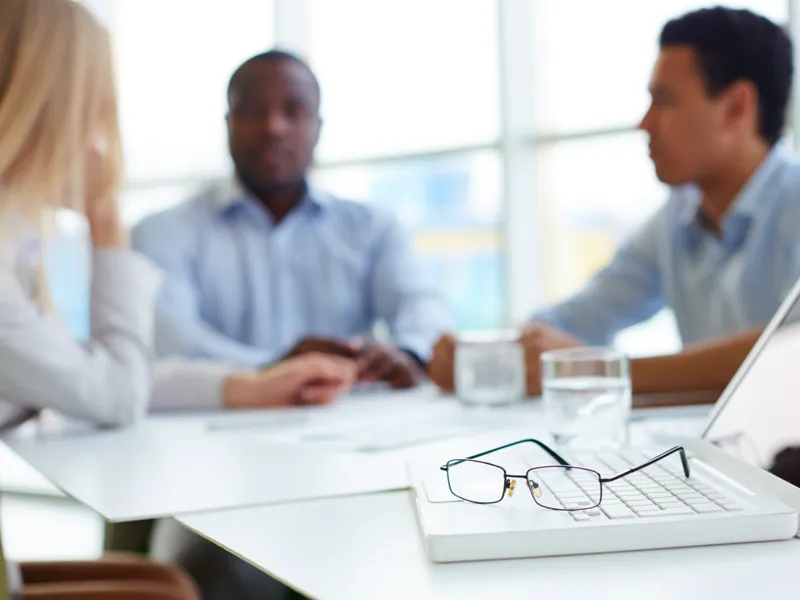 Glasses sit on a laptop computer as coworkers talk in the background