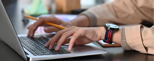 A person typing on a laptop computer while holding a pencil.