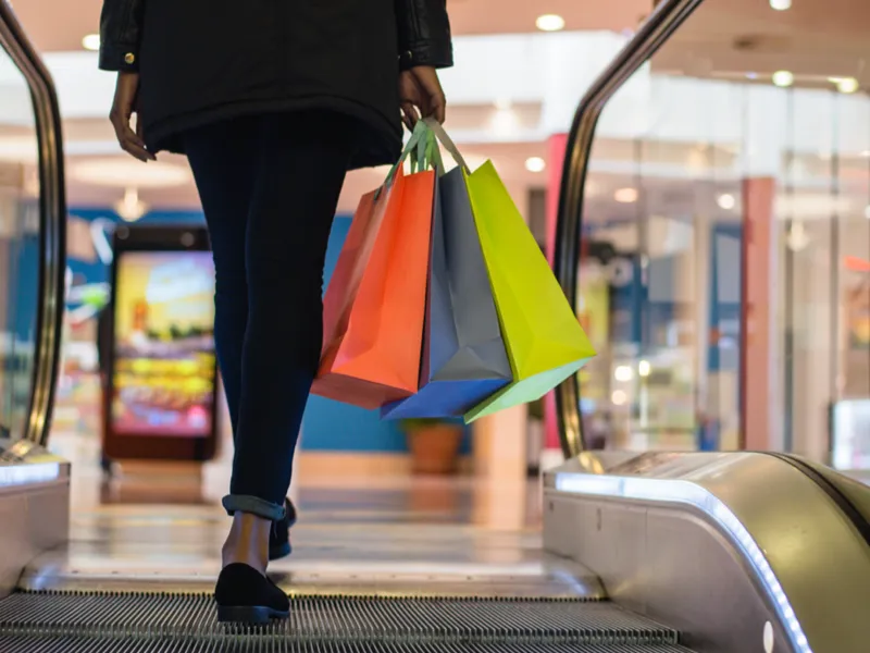 A person stepping of an escalator, holding three shopping bags.